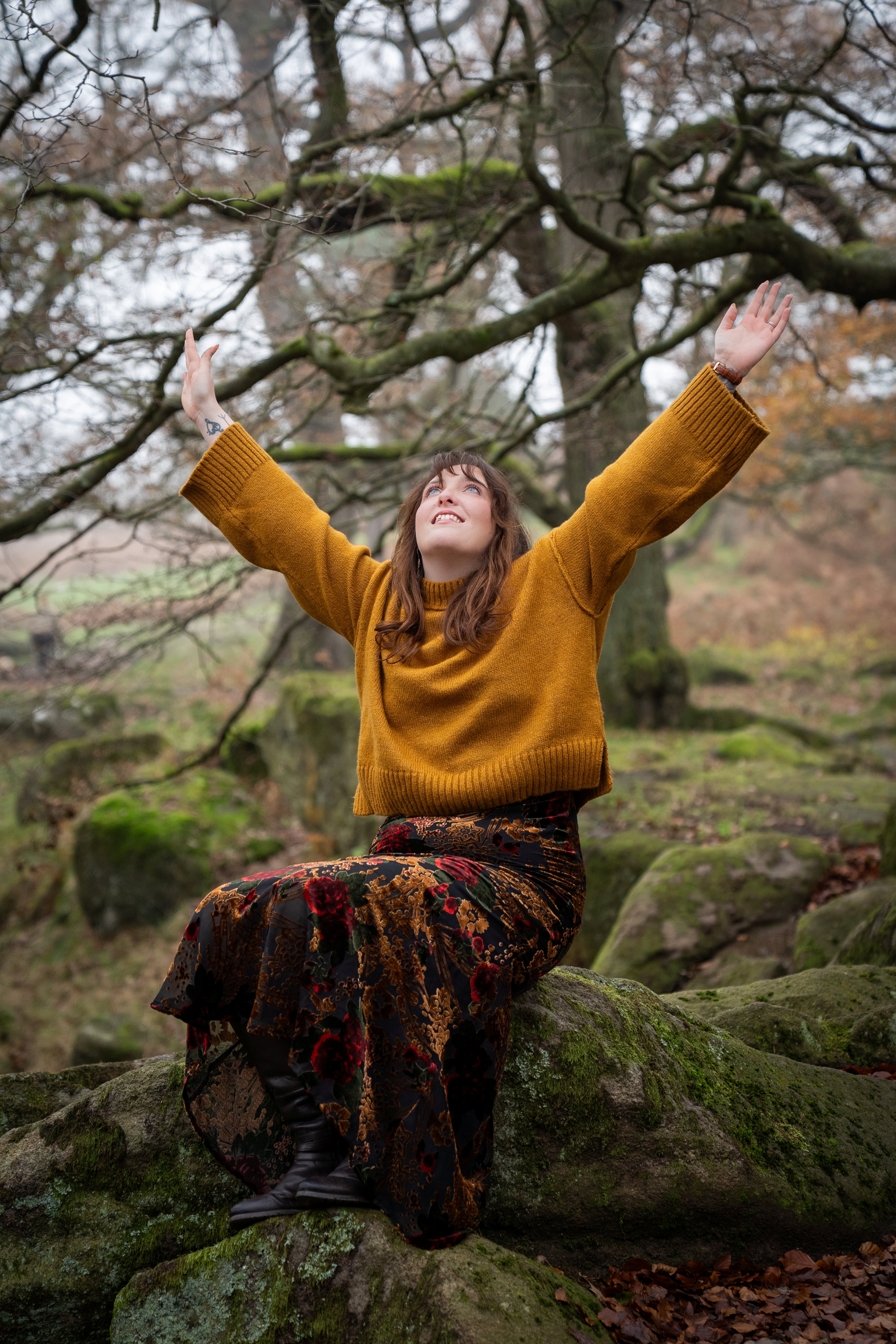 Nord-Essence founder Rhiannon sits on a moss-covered rock in a quiet forest, arms raised toward the sky. She wears a mustard yellow sweater and a dark floral skirt, surrounded by bare trees and fallen leaves — a moment of earthy stillness and connection.