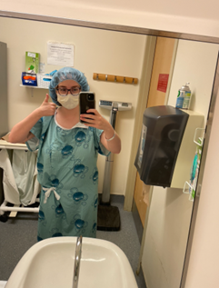 Photo of Madison in a hospital bathroom wearing a mask, shower cap, hospital gown. Taking a mirror selfie while holding a thumbs up
