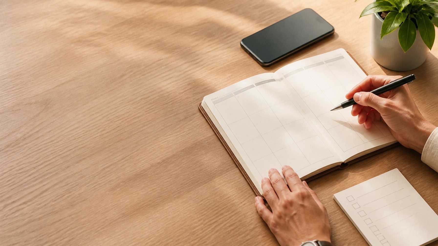 Person sitting at a sunlit wooden desk preparing to write in an open planner beside a small potted plant.