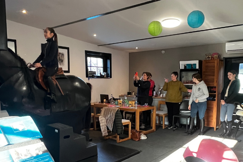 Ruiter op een paardrijsimulator tijdens een workshop over houding en zit, terwijl Nadia Molenaers en andere deelnemers meekijken