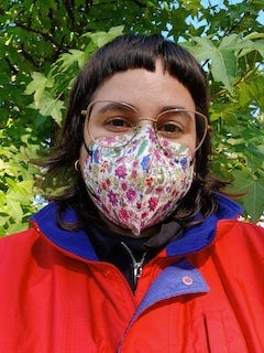 Photo de moi devant un arbre à feuilles vertes. Je porte un masque FFP2 à fleurs et une veste rouge.