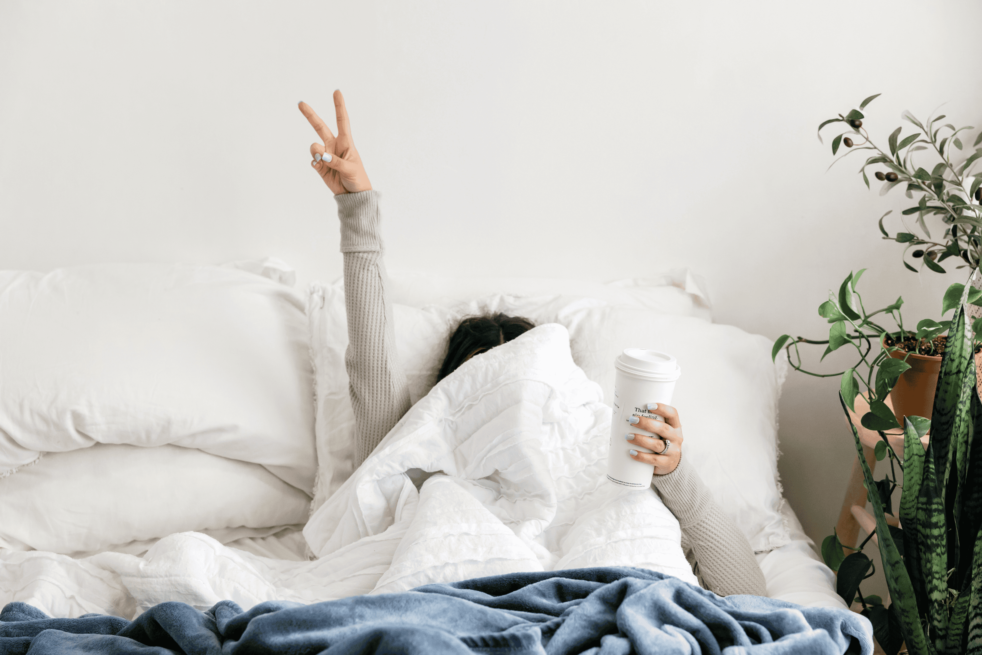 Femme dans son lit qui se cache sous les draps, un café à la main