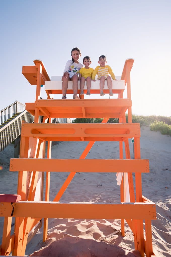 3 children sitting on an orange life guard stand on the beach. The sun is coming in behind them creating a backlit photograph.