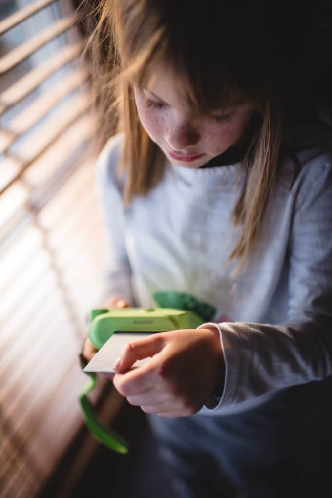 Child in front of window holding green camera printing off a polaroid. Showing photography for kids.