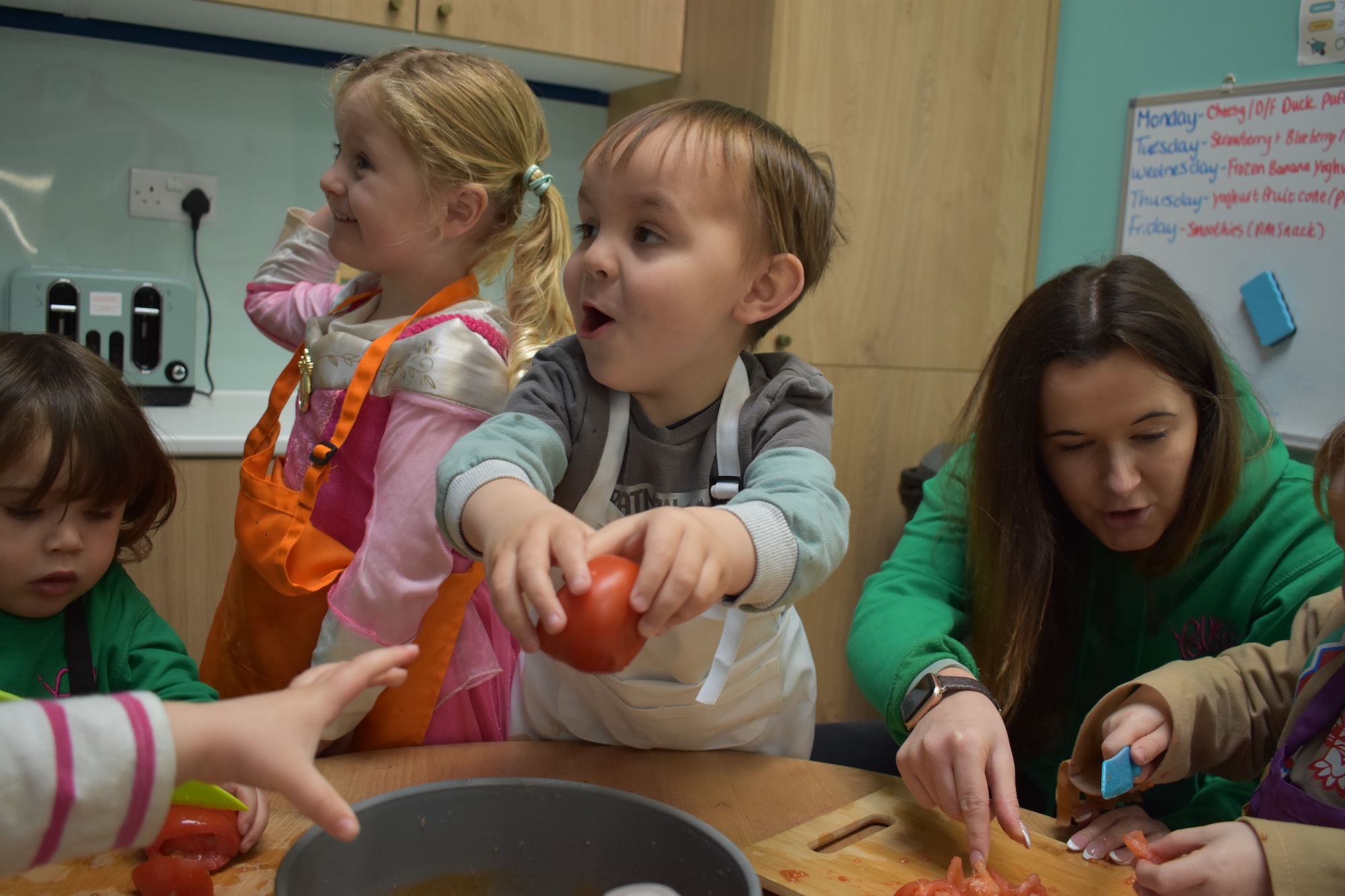 A woman teacher, with five children round a table, in an early years setting, with them learning about cutting tomatoes.