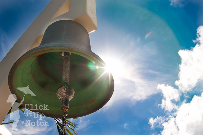 A bell partially blocking the sunny of a blue sky day with clouds in the sky creating a starburst photograph.