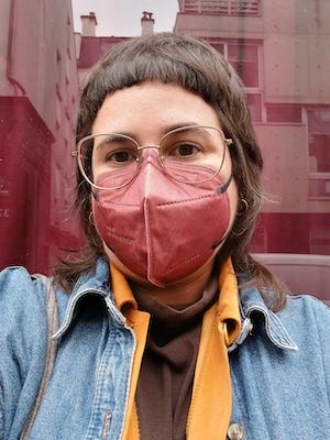 Photo de moi devant une devanture avec des rideaux bordeaux. Je suis vêtue aux couleurs de l'automne: je porte un masque FFP2 rouge bordeaux, un col roulé marron, et une chemise orange sous une veste en jean.
