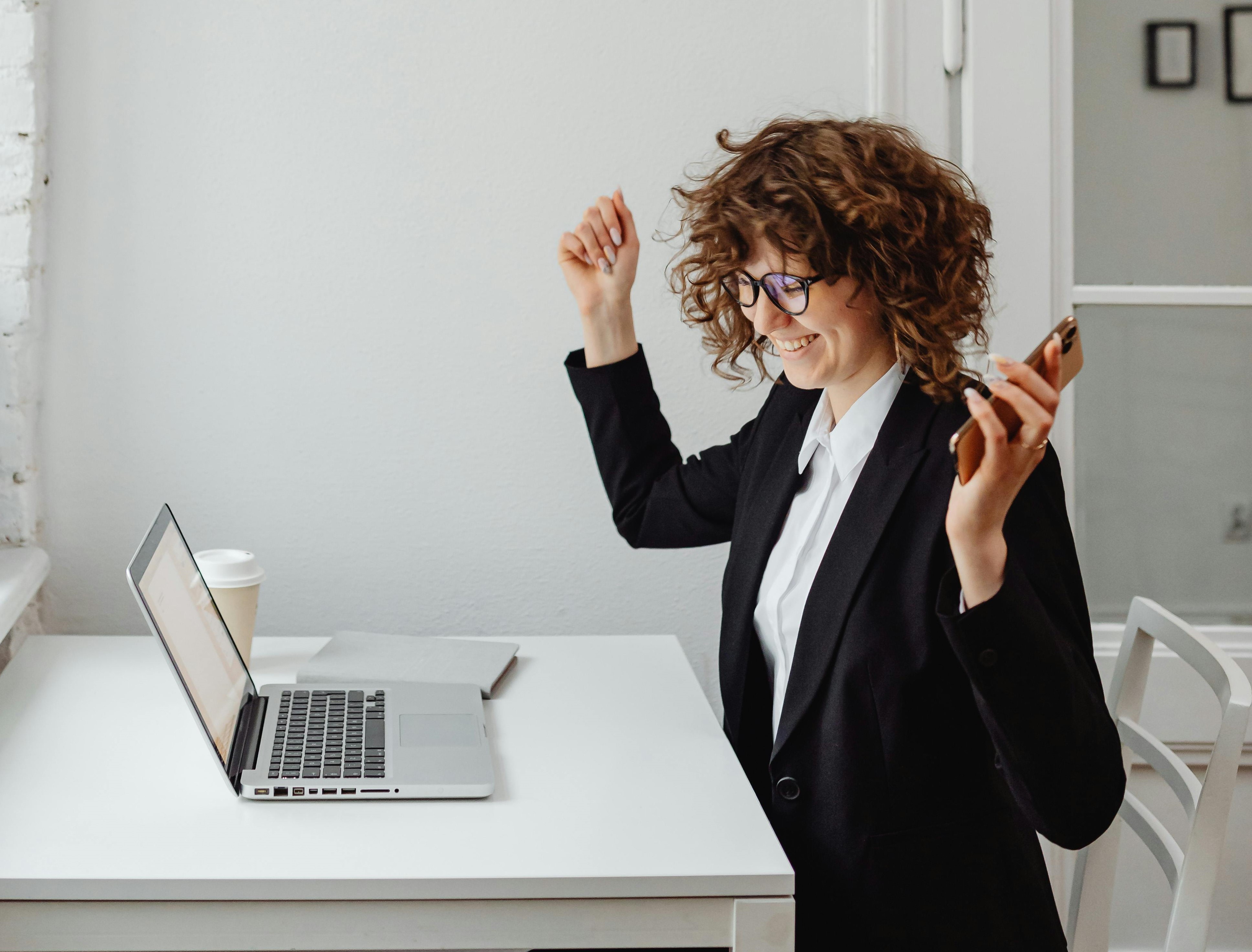 women smiling, sitting at desk with laptop, her arms up in the air in celebration