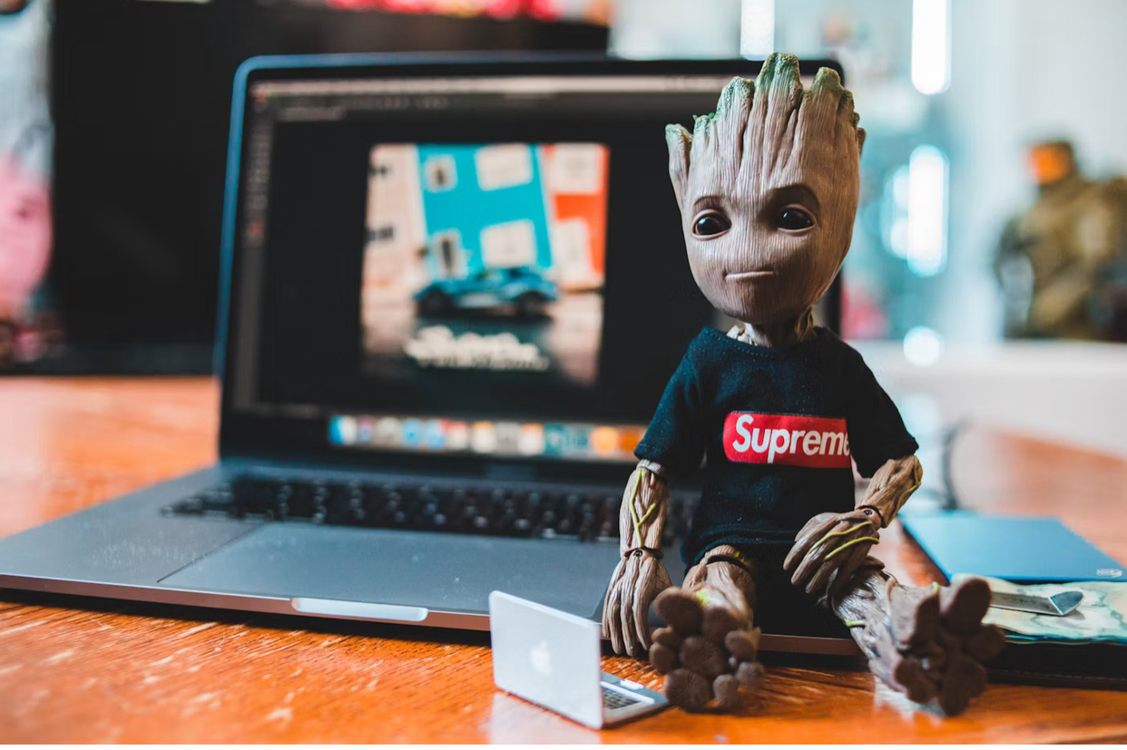 A picture of Groot of Marvel sitting next to a laptop with a shirt that reads Supreme