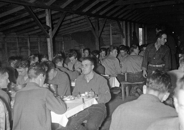 Dozens of soldiers sitting at eating at long tables in a mess hall.