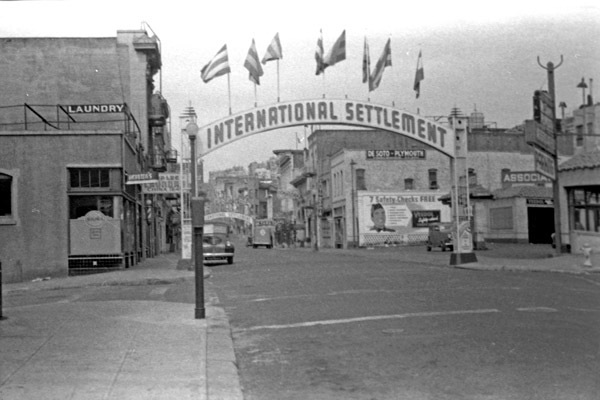 A city street with an arch that reads, "International Settlement" with numerous flags flying atop it. The street has a few cars parked along it.