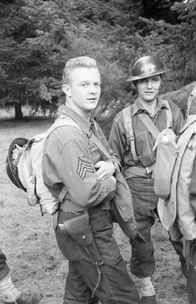 Two soldiers standing and looking at the camera.  The man on the left has their helmet off, while the one on the right is wearing their helmet.