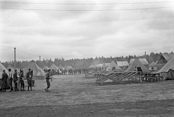 A small line of soldiers in front a camp of tents. A larger line of soldiers can be seen a few tent rows back in the background.