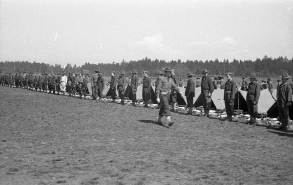 A line of soldiers standing with their backs to a line of tents, apparently preparing for inspection. In front of the tents are neatly organized groups of equipment, most likely the contents of the tent and the soldiers' belongings. A single man is walking along the line of standing soldiers.