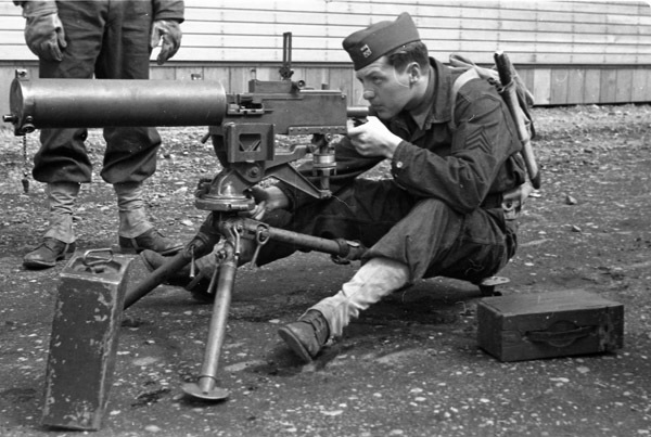 A soldier sitting on a seat behind a tripod-mounted machine gun.  He is looking through the sights of the gun.