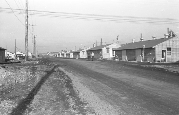 A line of single story houses on the right, with a dirt road on the left.  Two people can be seen walking down the road with their backs to the camera.