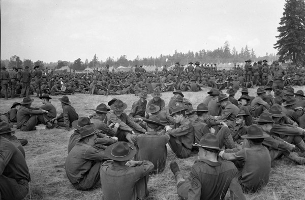 Hundreds of soldiers sitting or standing in relaxed positions in a field. Some fir tress are seen in the background.