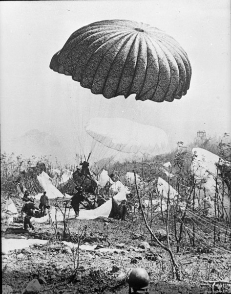 A paratrooper is just a foot off the ground and landing in his parachute. Multiple other soldiers are nearby on the ground with their chutes still attached, probably having just landed.