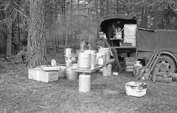 Three soldiers are organizing pots and boxes of food on a table and taking them off or on a military truck.  They are in a heavily wooded area.