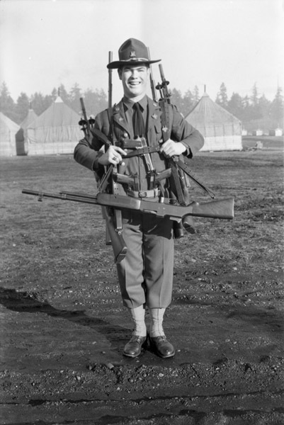 A smiling soldier equipped with multiple rifles, handguns, and swords, most slung over his shoulders and back. One rifle hangs from his neck, while two handguns are positioned on this belt.