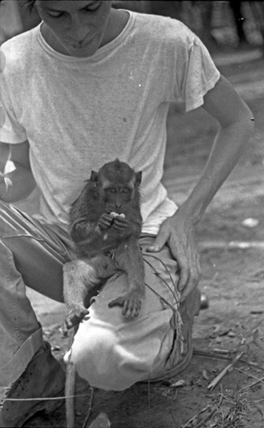 A monkey, named Jocko, sitting in the lap of a squatting young man. The monkey is eating something while looking toward the camera.
