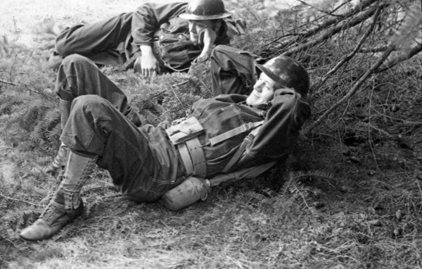 Two soldiers in full gear lay on the ground relaxing against some tree branches.