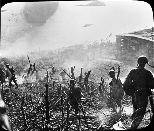 A half dozen soldiers walking down a hill away from the camera that has been cleared of most foliage, possibly by a battle. Smoke is seen throughout and a destroyed building is seen off to the right.