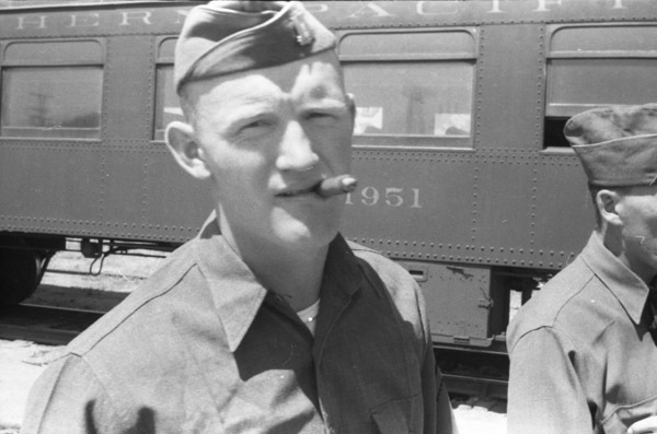 A soldier wearing a garrison cap and smoking a cigar facing the camera. A train car with "1951" printed on it can be seen in the background.