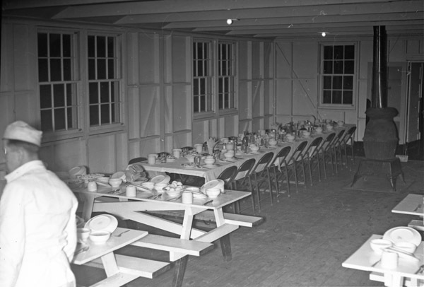 Tables in the mess hall with dirty dishes on them.  No one is at the tables.  One person is seen to the bottom left, facing away from the camera.