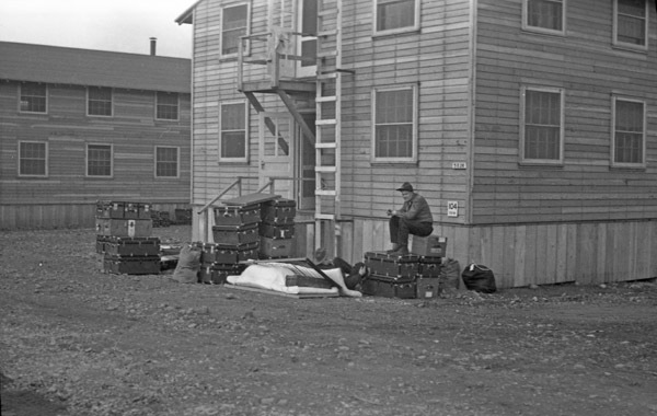 A soldier sitting on a stack of storage trunks outside of a barracks.  Other stacks of trucks are also seen.