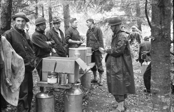 Four soldiers stand on the left of a table with pots and boxes of food. Two men stand to the right, the closest wearing a rain poncho.