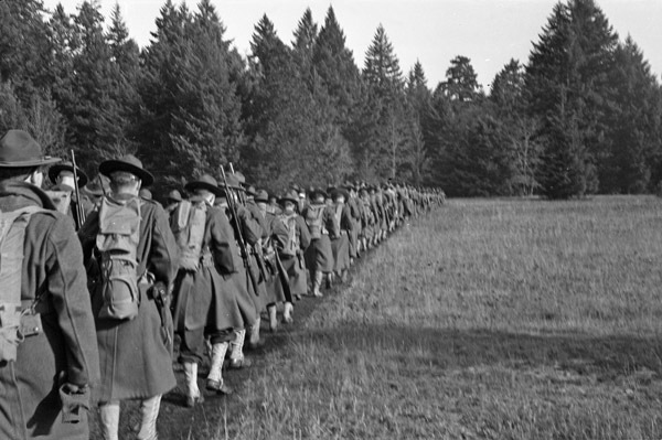 Dozens of soldiers marching two or three abreast in a line through a wooded clearing. They are marching towards the tree line.
