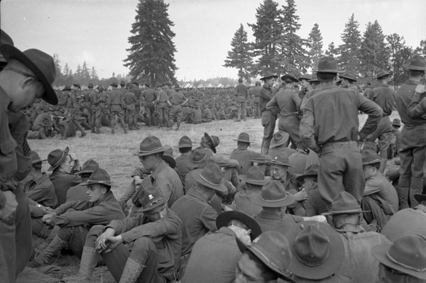 Hundreds of soldiers sitting or standing in relaxed positions in a field.  Some fir tress are seen in the background.