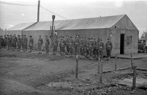 Around two dozen soldiers in uniform stand at attention with their backs to a building.