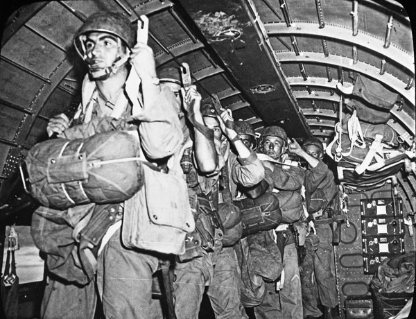 A half dozen soldiers in full paratrooper gear standing in the interior of an airplane holding to hooks that secures them in place.  They look to be getting ready to parachute out of the plane.