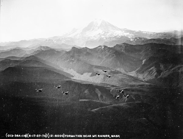 Nine airplanes, flying in three triangle formations, flying over mountains.