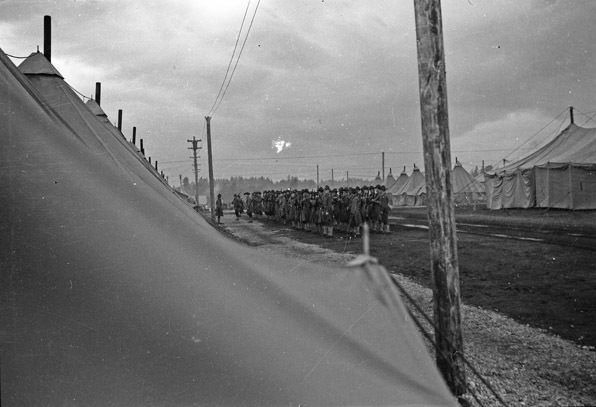 Rows of dozens of soldiers standing in formation in a large path surrounded by tents.  An officer is facing the formation.