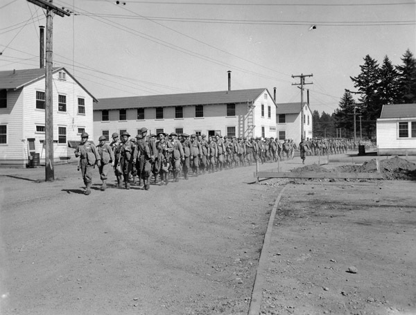 Dozens of soldiers walking in a line, three-to-four people wide, past a series of two-story buildings on their right, and single-story buildings on their left.