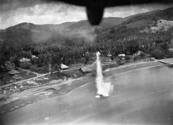 A plume of water showing a bomb exploding in the water near a shoreline populated by houses and other buildings.