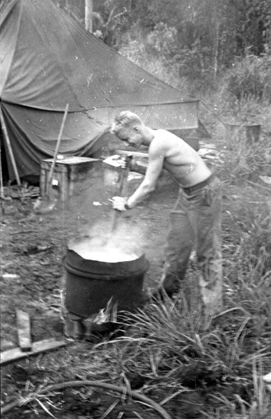 A shirtless soldier uses a pole to to stir clothes in a steaming bucket.  A tent and trees are in the background.
