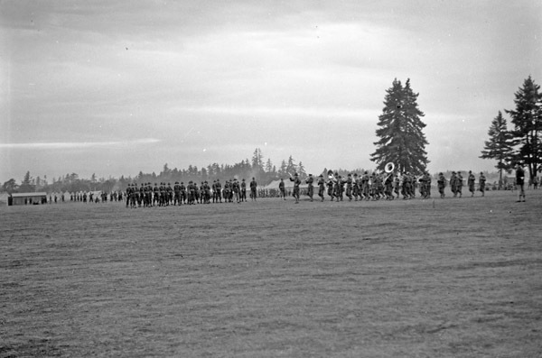 A line of soldiers playing musical instruments in a parade formation marching. More soldiers and tents can be seen in the background.