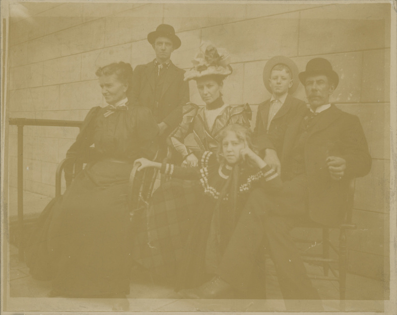 Three women, a man, sit for a posed photo, while two young boys stand in the background.