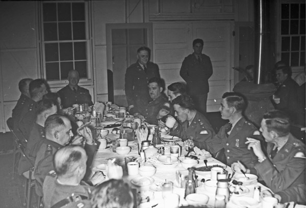 A group of soldiers sitting at a long table having a dinner.  While some are standing, most are sitting while eating and drinking.