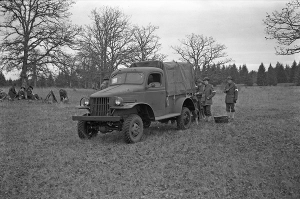 A military truck in a wooded clearing, facing just the the left of the camera.  A few soldiers are standing near the back of the truck, while a few others are sitting down near a tree to the left of the truck. 