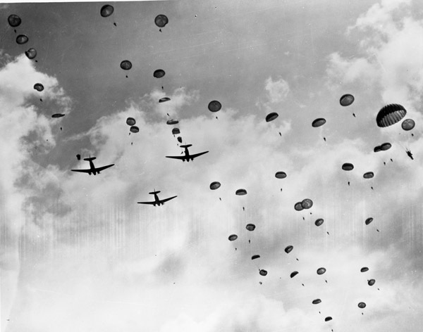 Three military planes in the sky flying away as seen from below. There are numerous paratroopers in the sky behind the planes in various stages of deploying their parachutes.