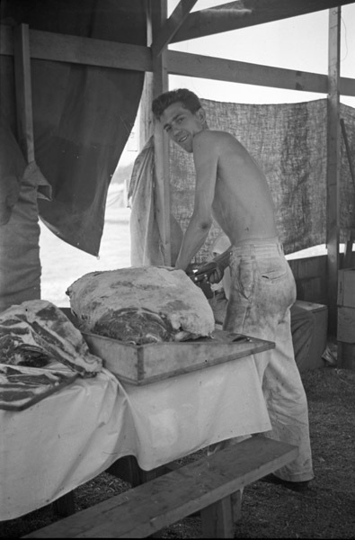 A shirtless man in a tent standing and looking at the camera over his shoulder while cutting a large slab of beef that is on the table beside him.