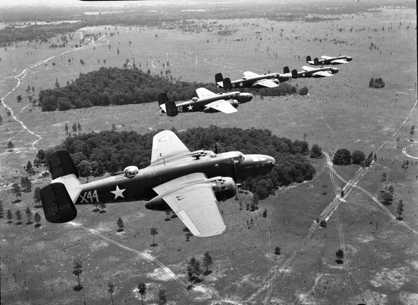 Five bombing planes flying over a sparsely forested grasslands, from the view of another airplane.