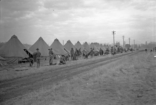 A long row of large tents along a dirt road.  Dozens of soldiers are standing or kneeling outside the tents, looking to be working.