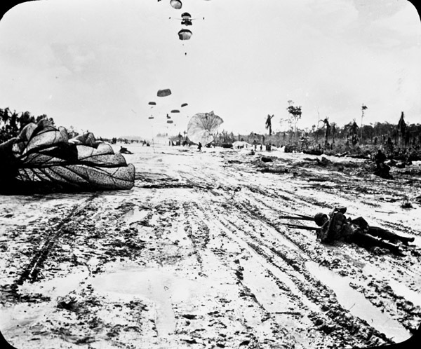 A soldier laying in the mud being pulled by his parachute. Many more paratroopers can be seen still landing in the background.