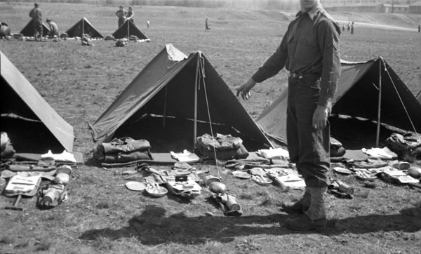 A soldier stands in front of a tent, where a large number of items, presumably the soldier's equipment, are arranged in front of the tent. Tents can be seen on either side of this one, as well as behind it, with similar equipment organized in front of them.